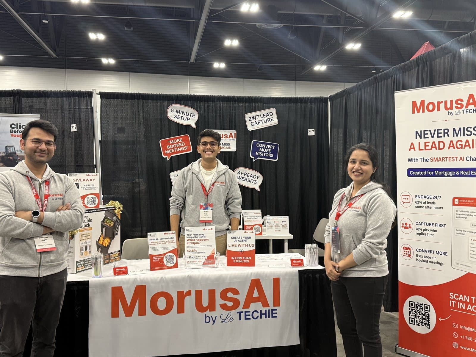 Three members of the MorusAI team in MorusAI hoodies at the MorusAI booth at RAECON 2026 in Edmonton, with a branded table, the giveaway air fryer, speech-bubble signs reading 5-minute setup, 24/7 lead capture and AI-ready website, and a Never Miss a Lead Again banner
